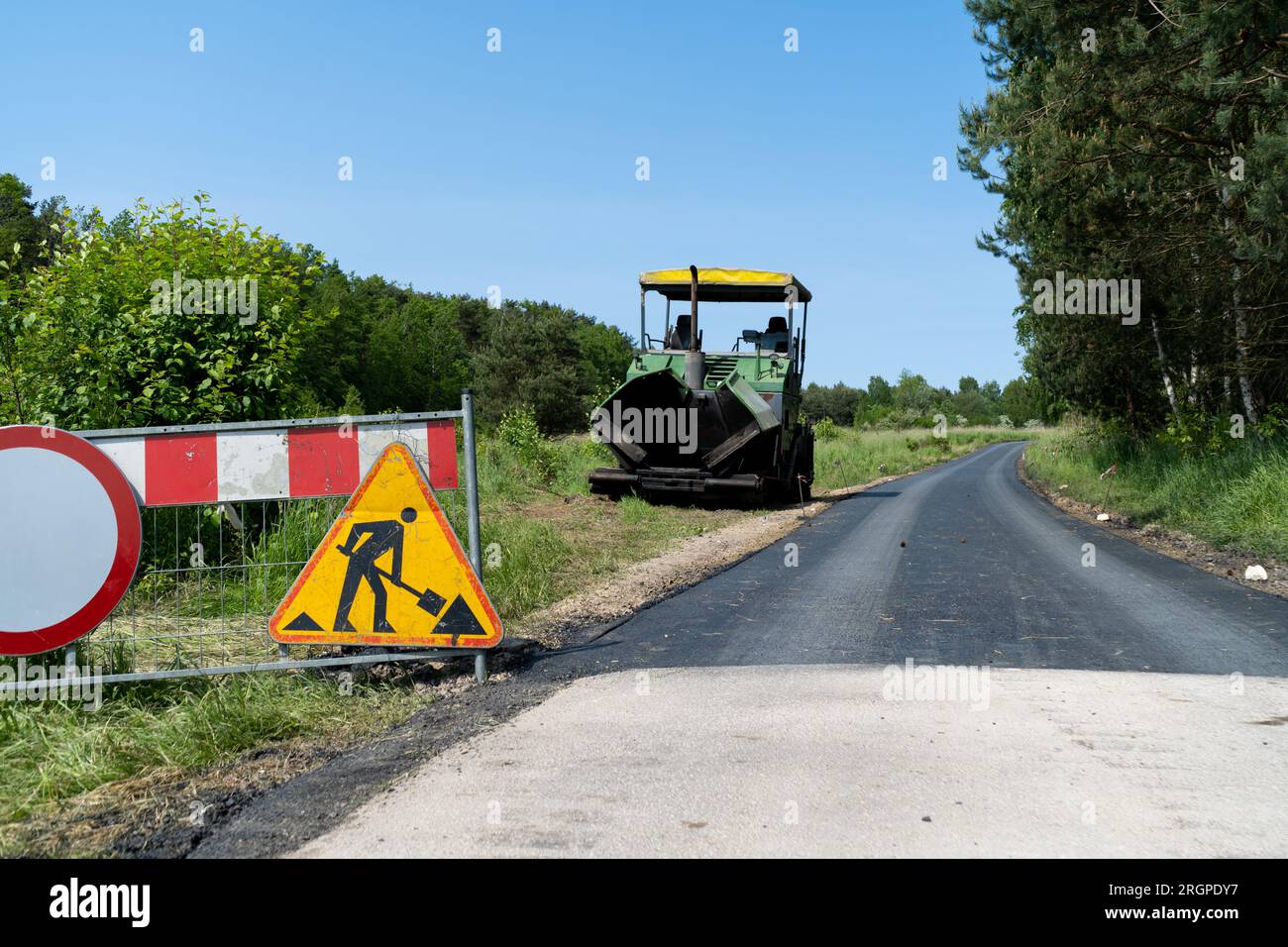 Road work ahead warning sign on a fence of new road construction site. Paver finisher equipment in bg. Crawler paving machine for laying asphalt. Stock Photo