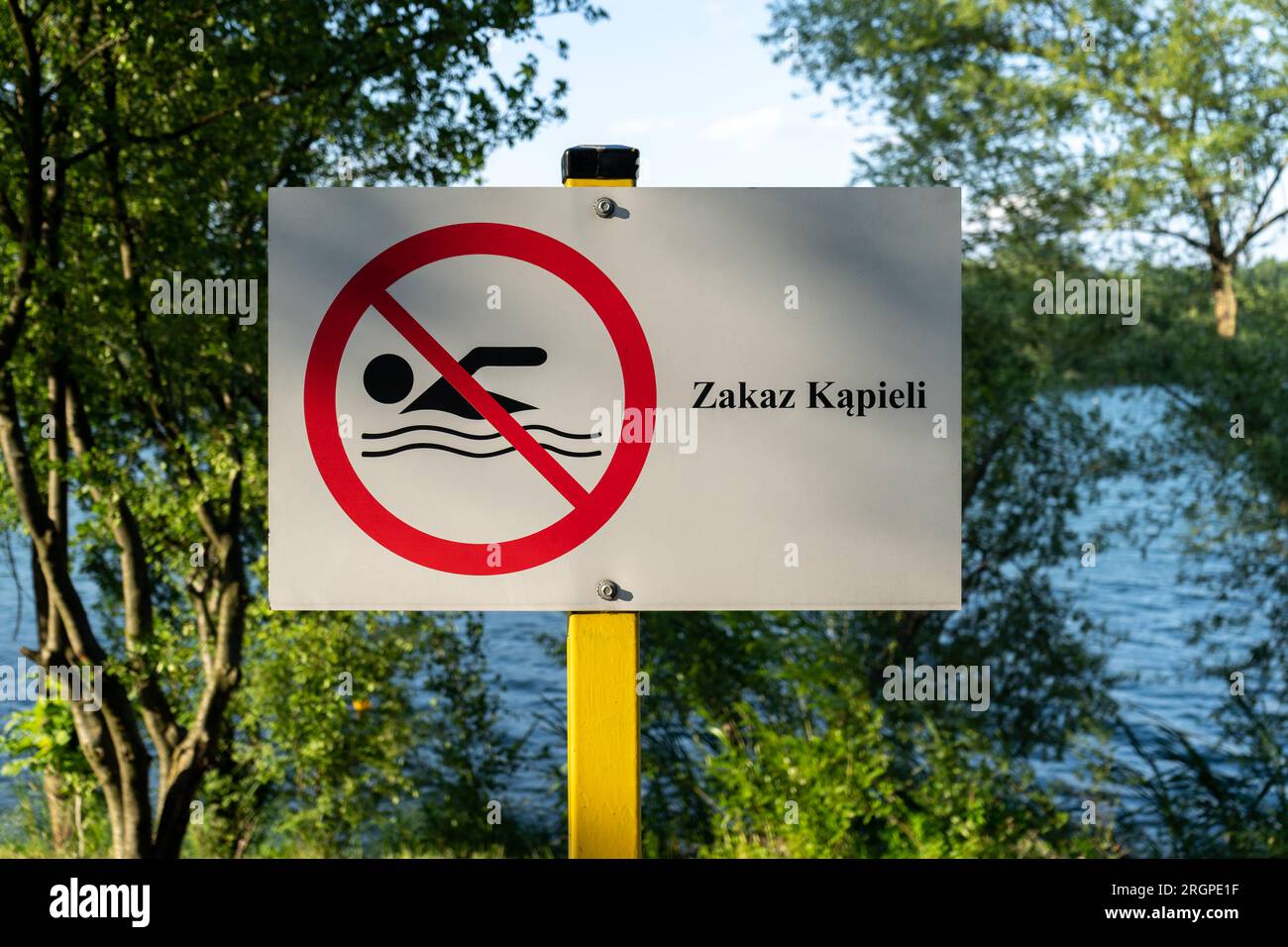 No Swimming sign post plaque. No swim area signage, mounted on water shore of lake in Poland. Text in Polish Zakaz kąpieli means Swimming prohibited. Stock Photo