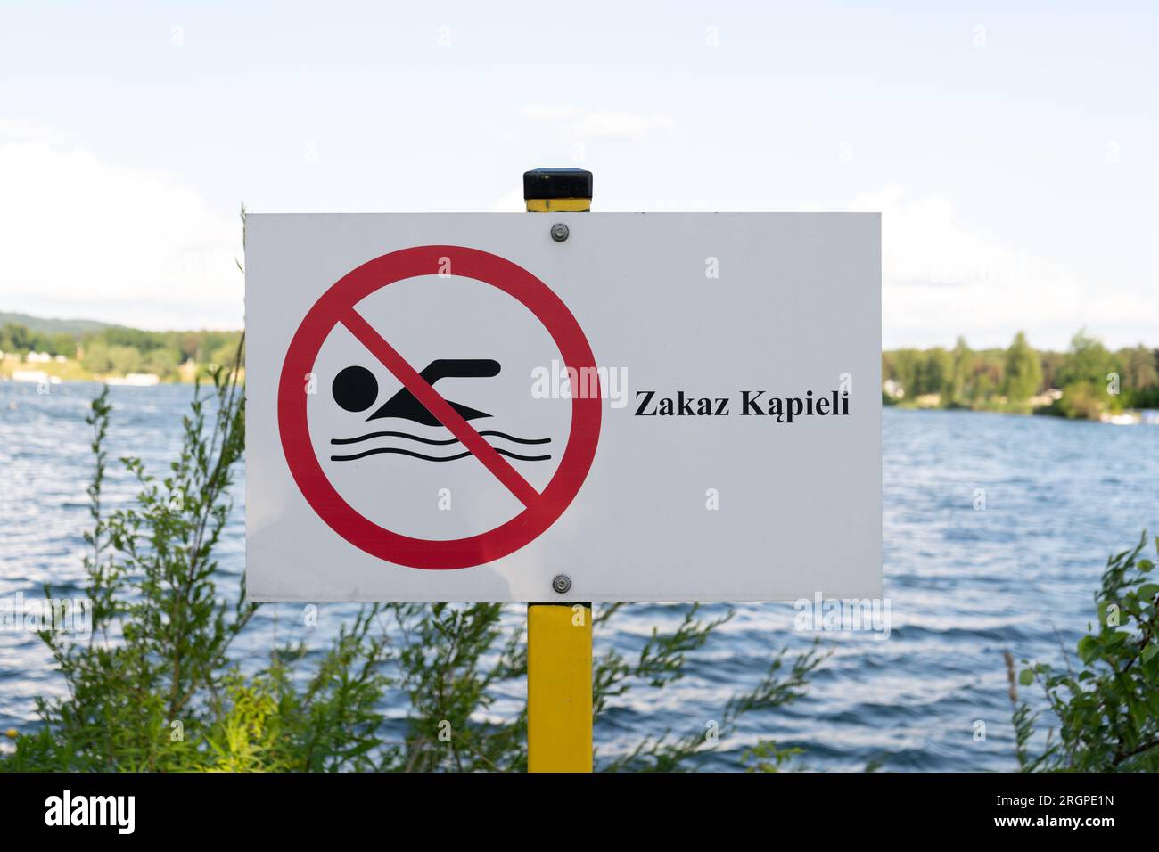 No Swimming sign post plaque. No swim area signage, mounted on water shore of lake in Poland. Text in Polish Zakaz kąpieli means Swimming prohibited. Stock Photo