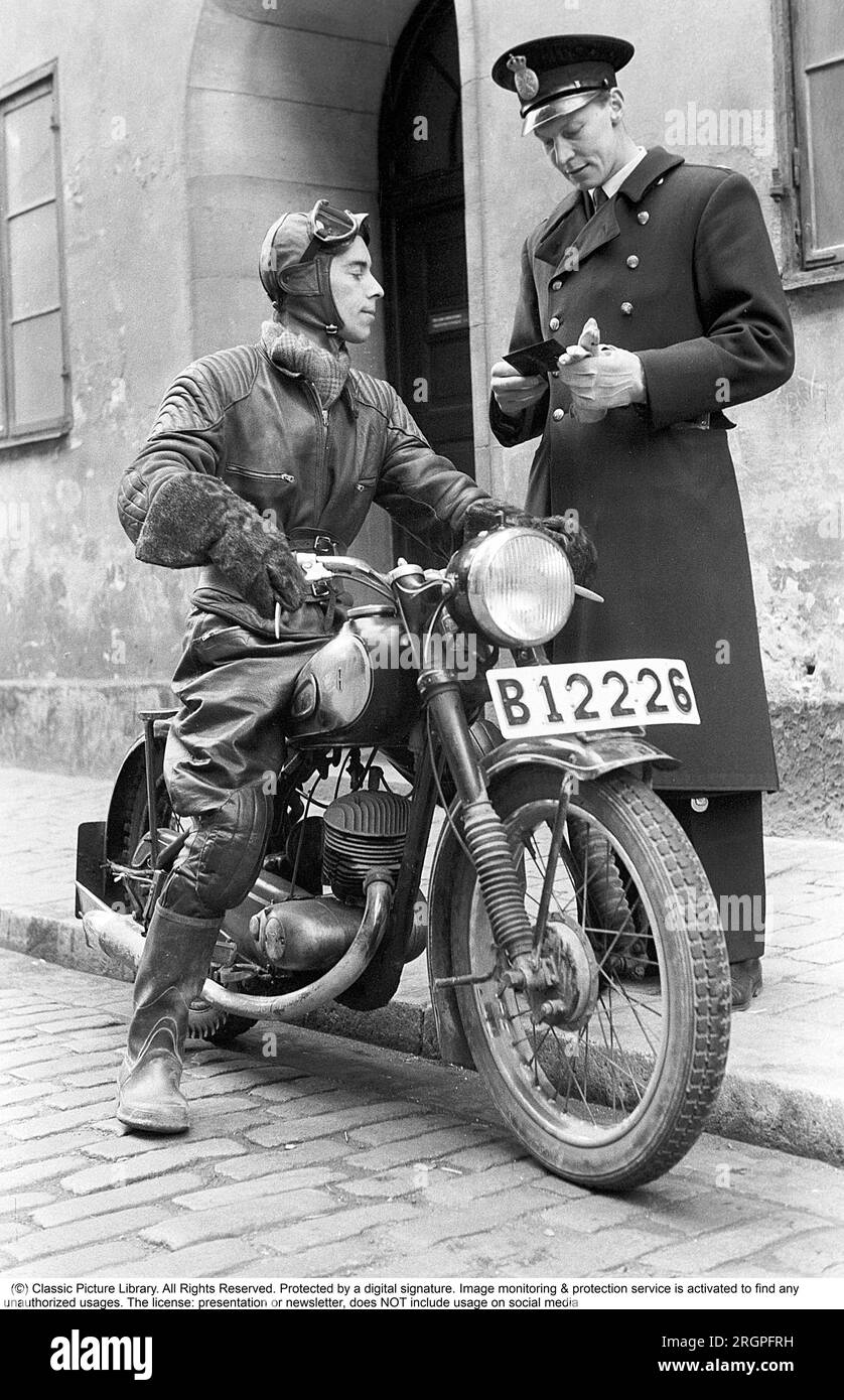 Motorcyclist in the 1950s. A young man dressed in the typical way the motorcyclists did in the 1950s. All leather. Leather boots, trousers and jacket. A kidney belt outside the jacket. For some reason a policeman is standing by his side on the sidewalk looking at what seems to be the motorcyclists driving license.  Sweden 1955. He is actor Sven-Eric Gamble, 1924-1976. Kristoffersson ref 2B Stock Photo