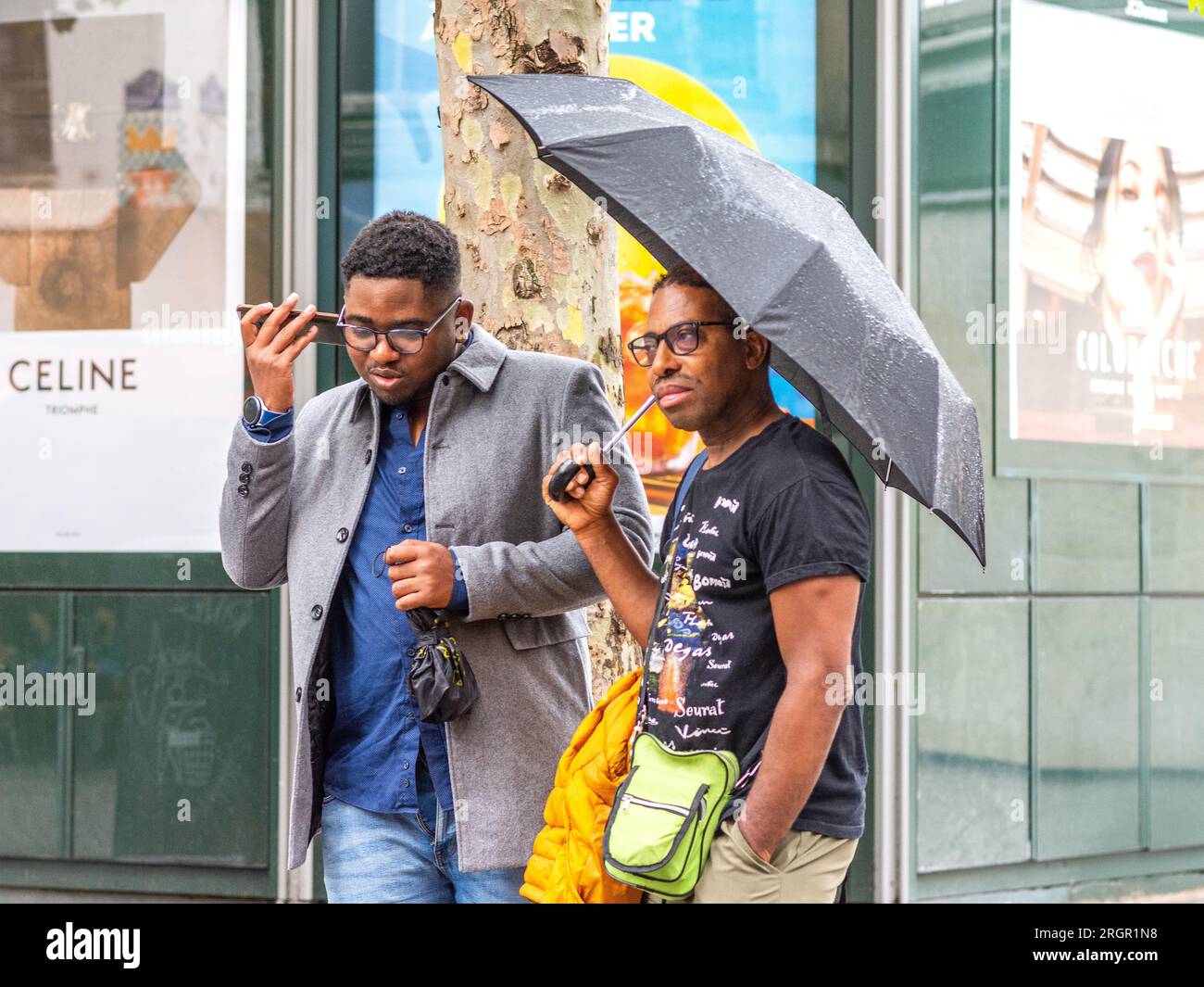 Two black male friends, one holding umbrella the other making phone call in rainy Paris, France. Stock Photo