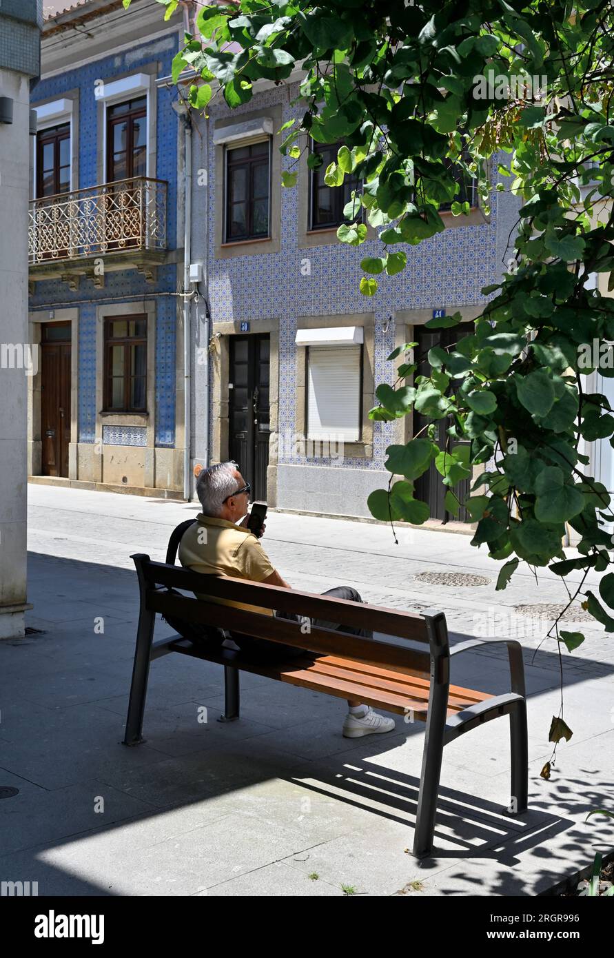 Older man sitting on bench relaxing in shade with traditional tilled building in background, Portugal Stock Photo