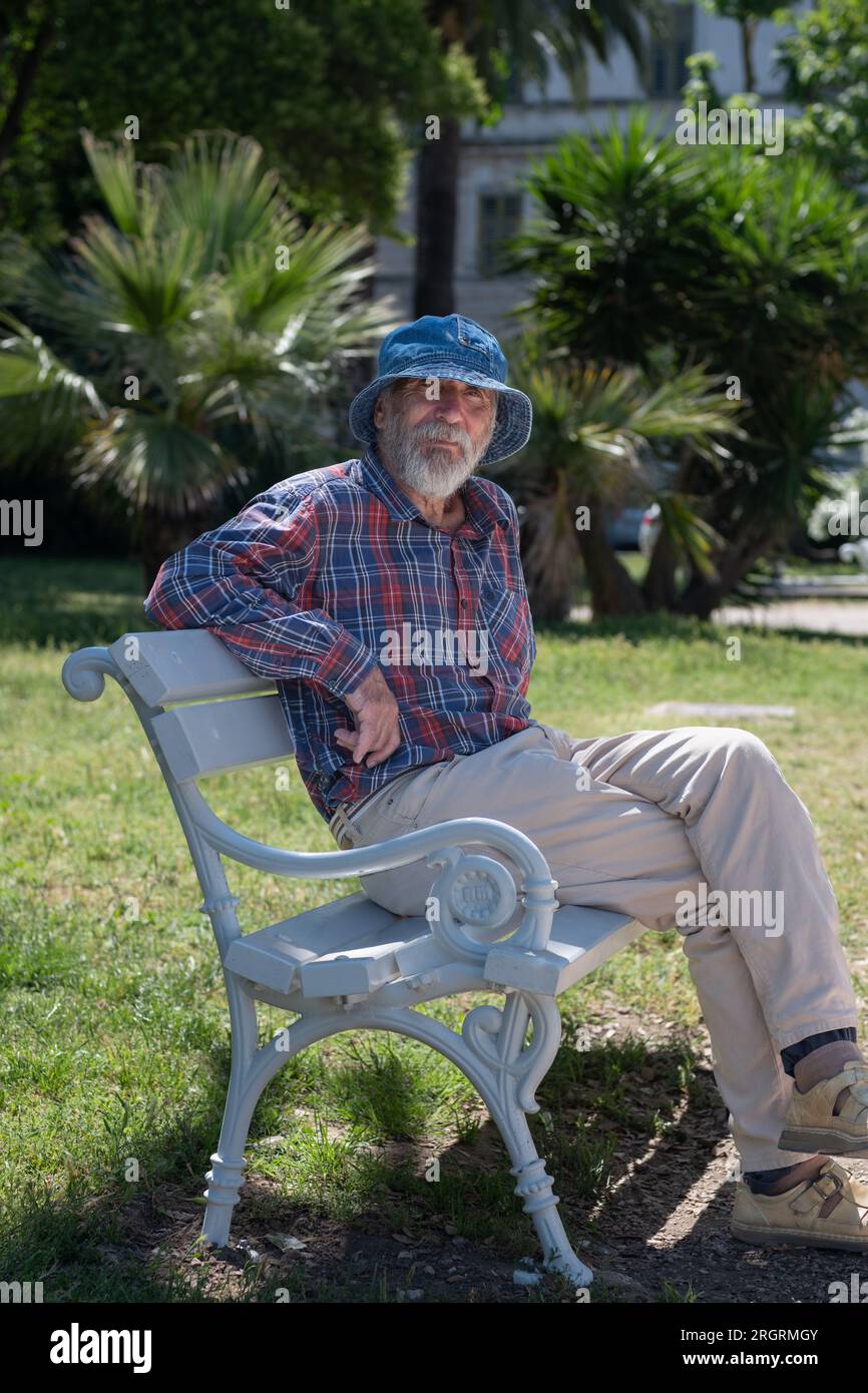 An old man with a beard in a panama hat and a plaid shirt sits on a park bench on a summer sunny day. Stock Photo