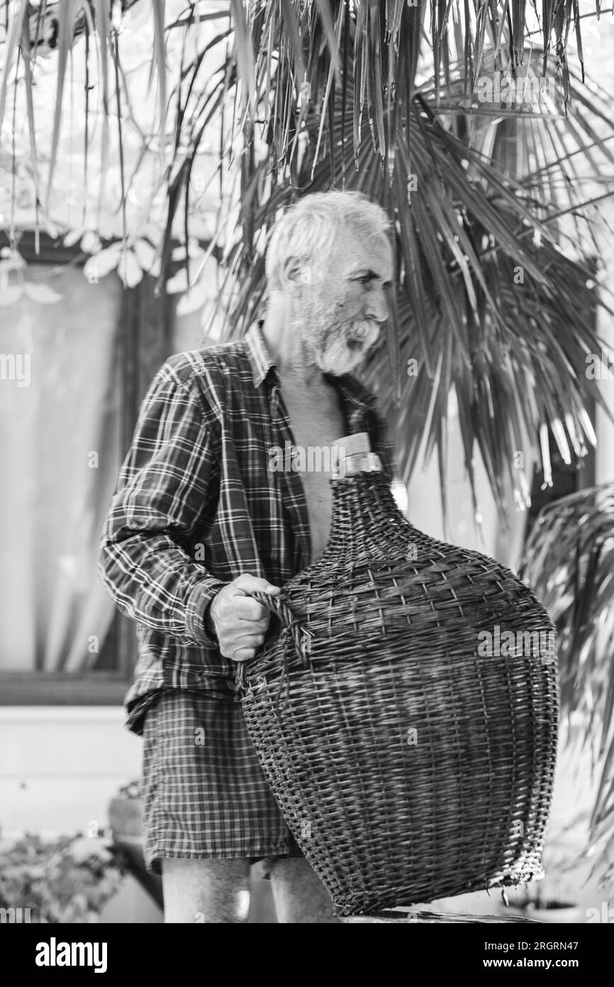 An old winemaker poses with a vintage big wine bottle wrapped in rattan under a palm tree. Charismatic handsome grandfather holds a bottle entwined wi Stock Photo