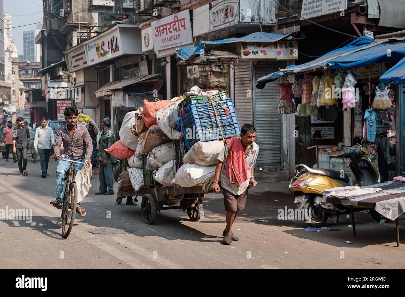 In a street in commercial area Bhuleshwar, Mumbai, India, a handcart puller struggles to pull an overloaded cart, while being overtaken by a cyclist Stock Photo