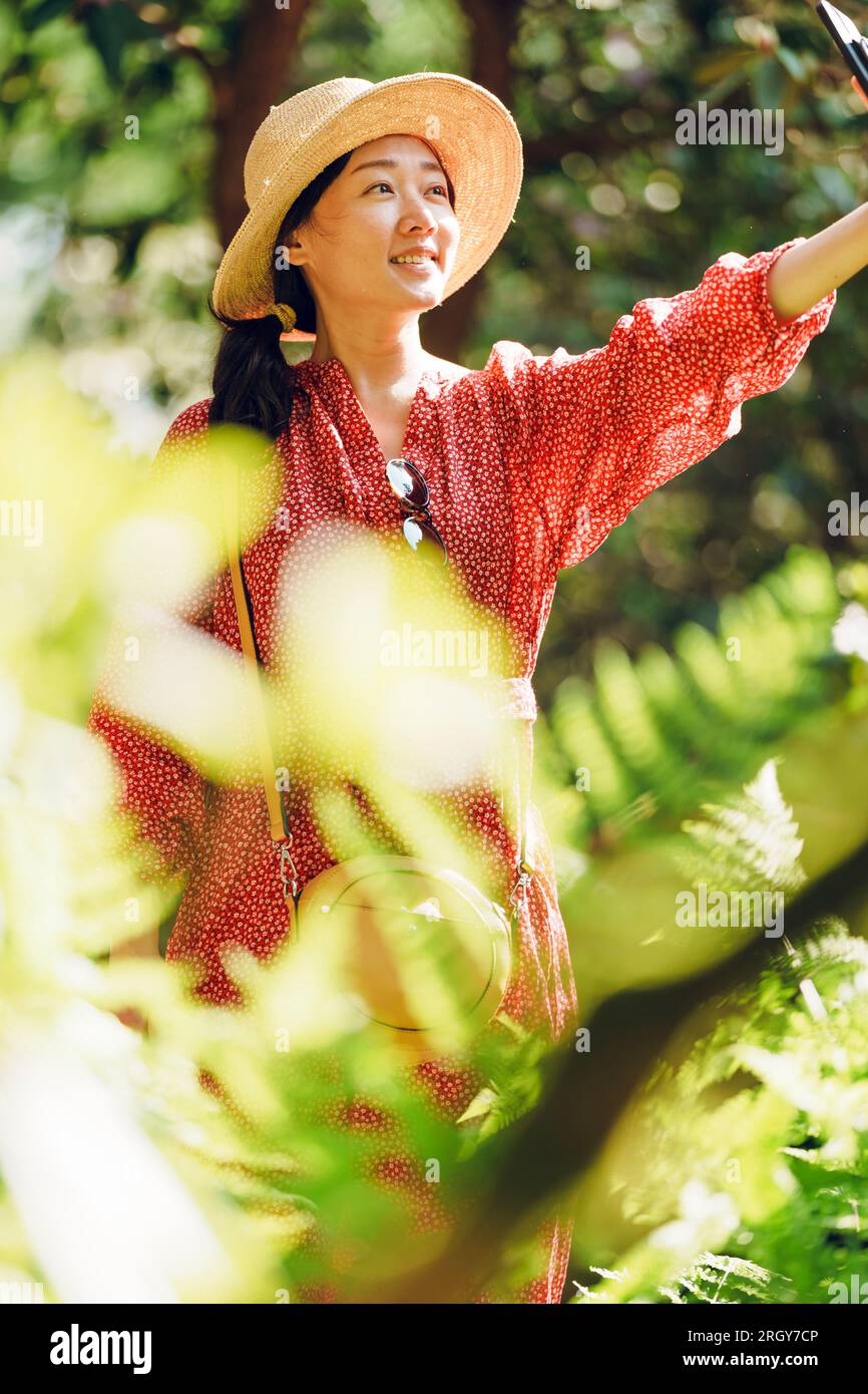 A woman taking selfie in a garden. She wears a read dress and a summer hat. Stock Photo