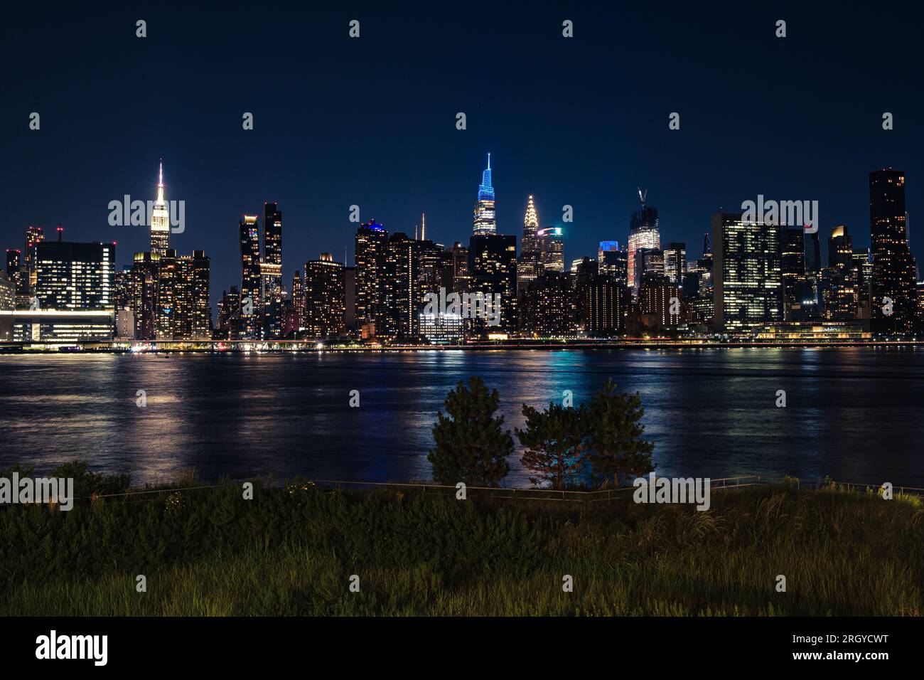 Iconic Manhattan skyline seen from Long Island City. Trees in the foreground, city lights on the cloudless summer night. Stock Photo