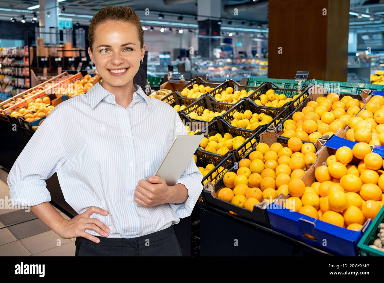 Young woman food retail store manager standing in the sale floor in front of boxes with fruits, lemons and oranges. Stock Photo