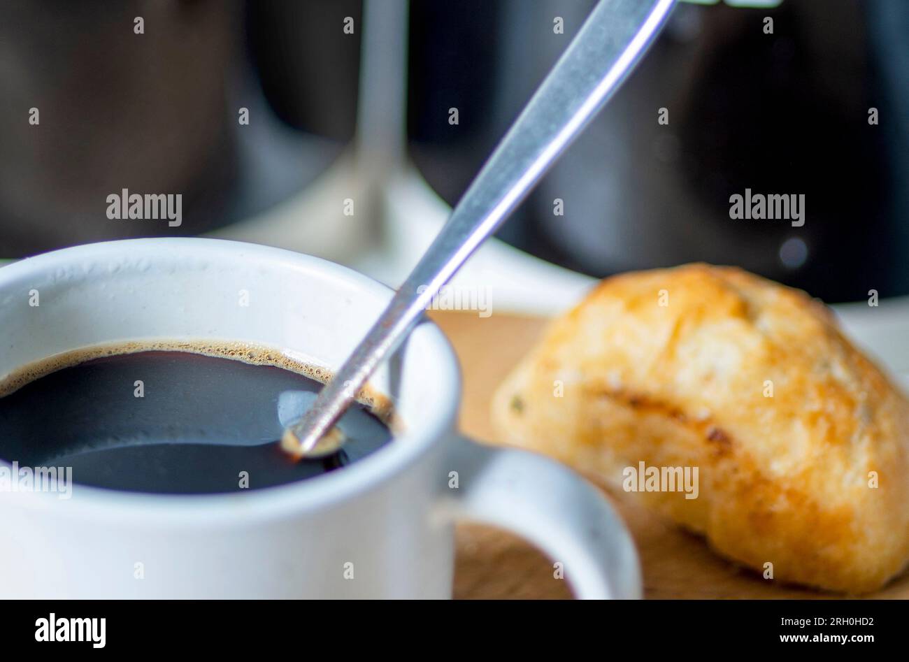 Freshly poured,hot,ready to drink,in an espresso coffee mug and silver spoon used to stir,on a wooden chopping board next to a bread roll,sugar and co Stock Photo