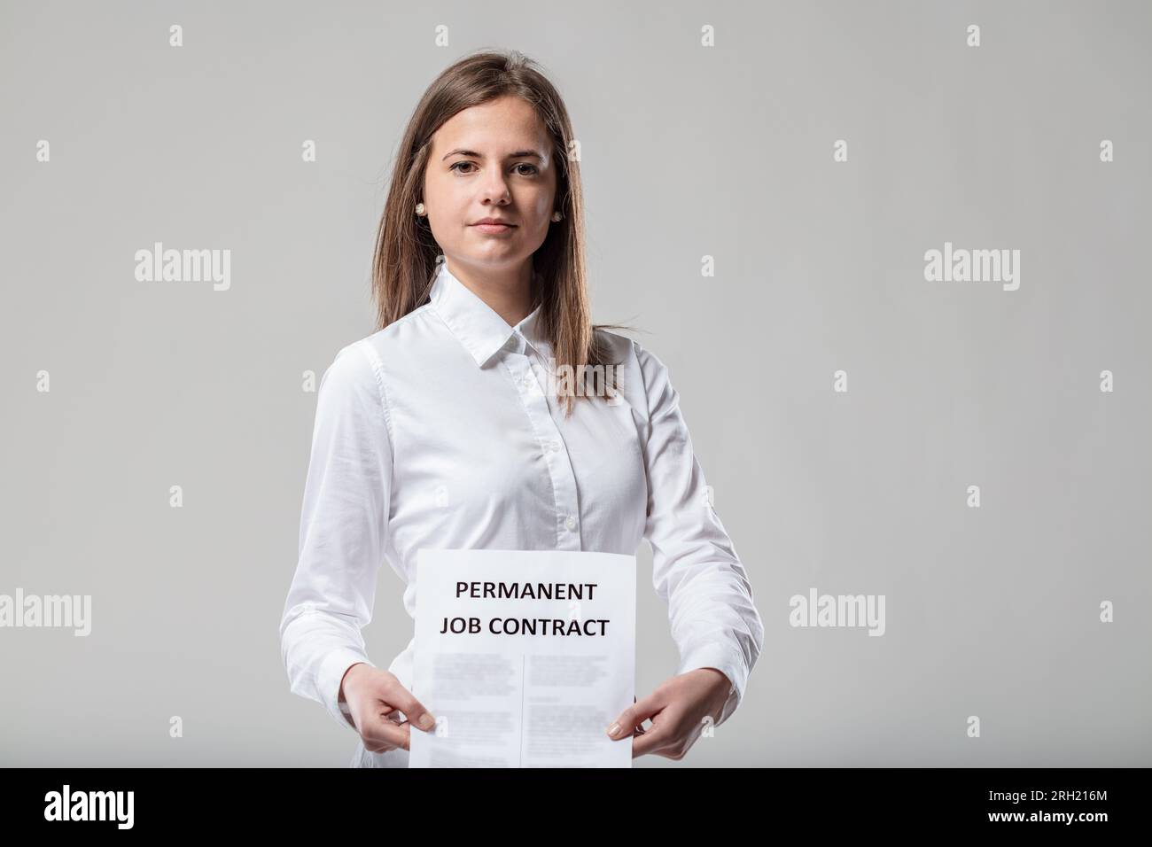 Woman in white shirt, proud expression, shows permanent work contract. Medium-length brown hair; importance of employee stability, business continuity Stock Photo