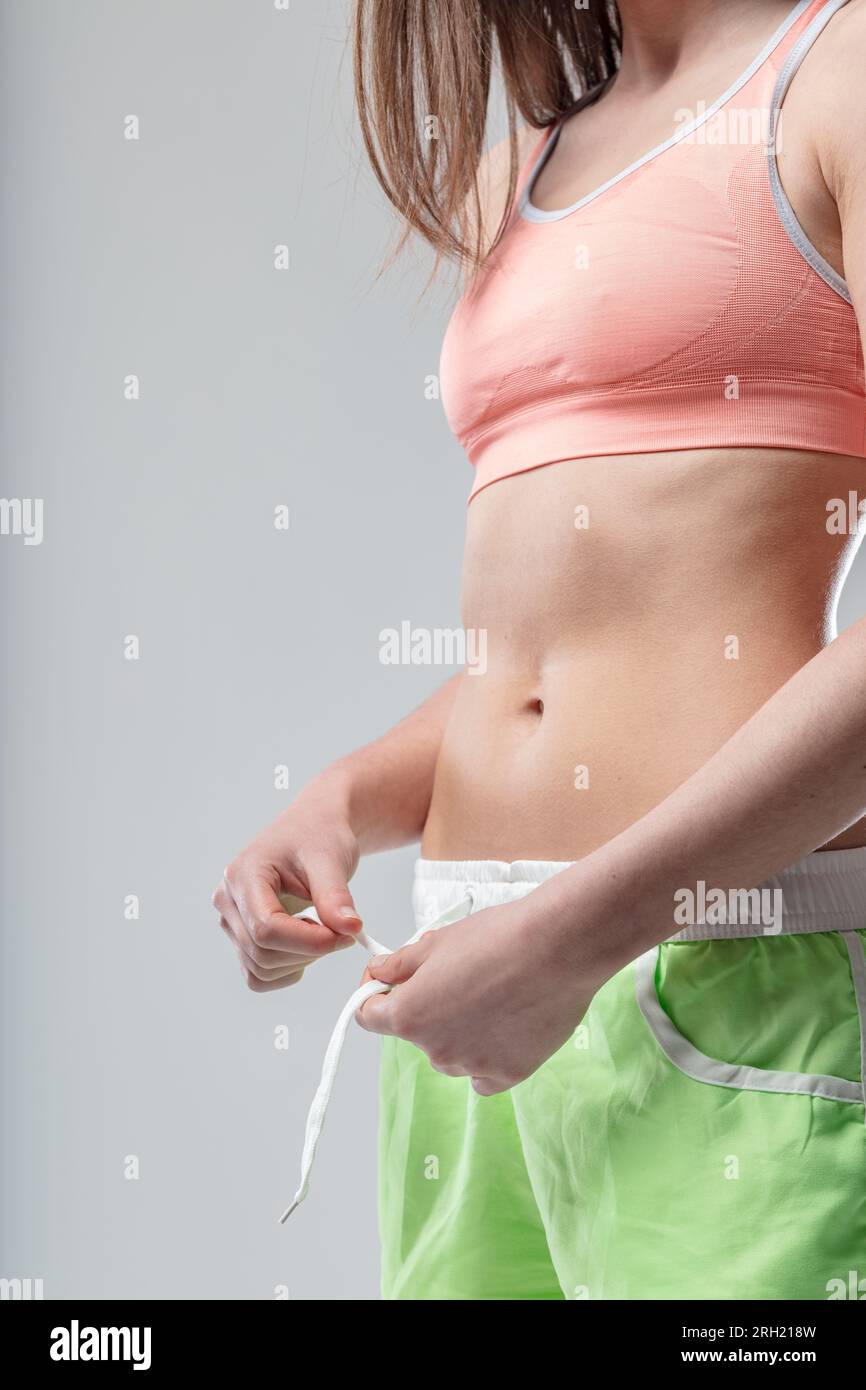 Flat stomach in focus, a fit young woman tying shorts, enviable midriff. Top or bra is orange, bottom part green. Face not visible, some hair seen Stock Photo