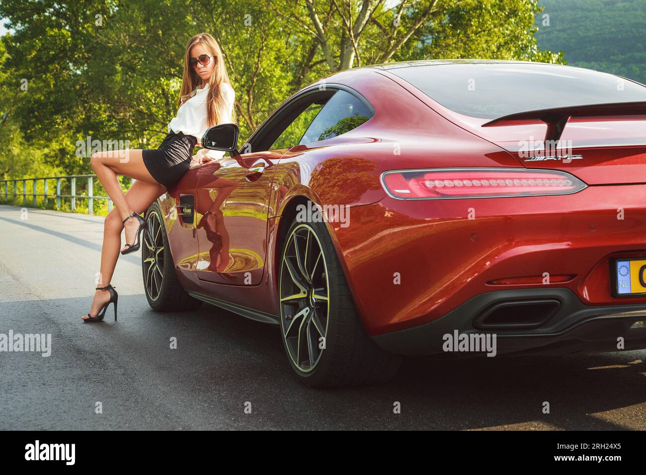 Red Mercedes AMG-GTS sportscar (2015) parked on a road with stylish female, wearing sunglasses, short skirt & high heels, leaning against the car Stock Photo