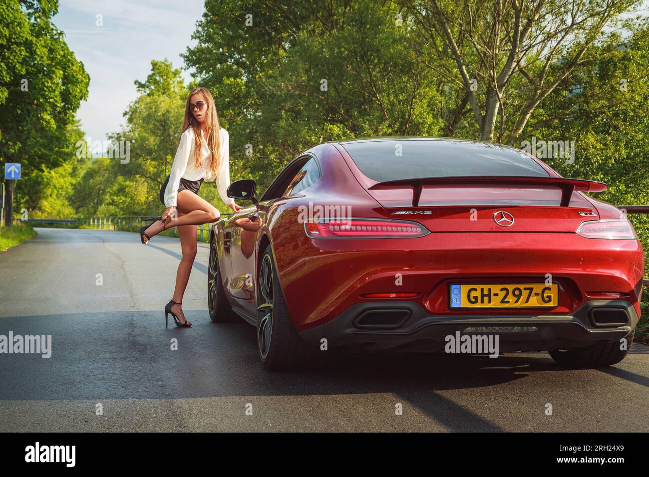 Red Mercedes AMG-GTS sportscar (2015) parked on a road with stylish female, wearing short skirt & high heels, leaning on car while adjusting her shoe Stock Photo