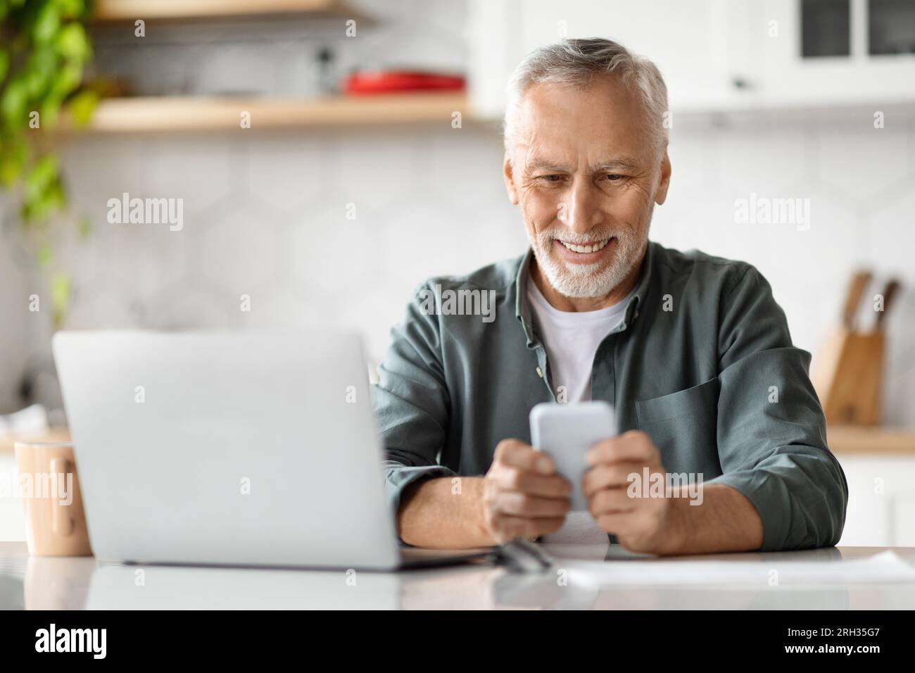 Smiling Senior Man Using Smartphone And Laptop Computer At Home Stock Photo