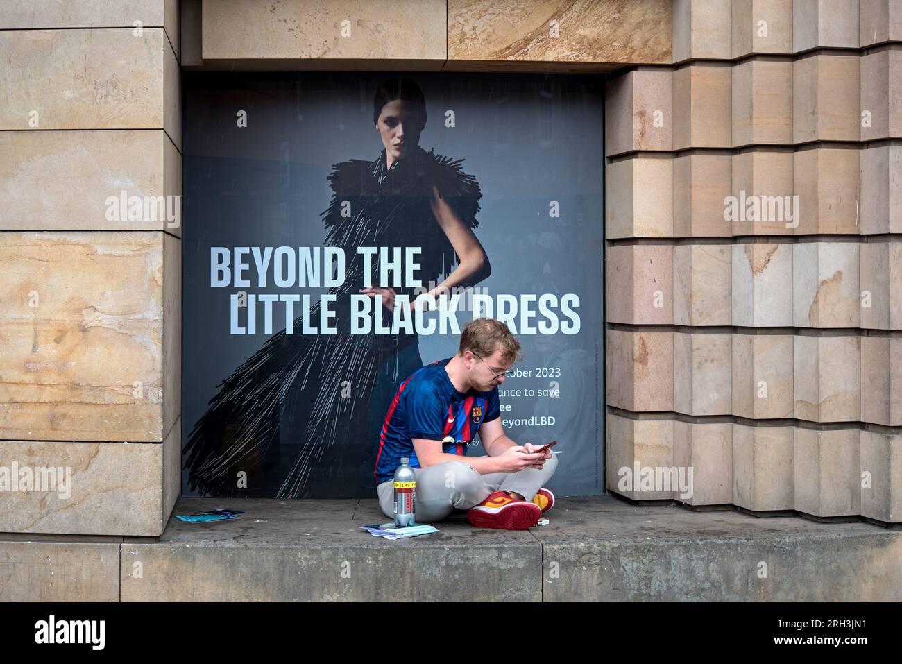 Young man checking his phone outside the Museum of Scotland in front of an advert for 'Beyond The Little Black Dress' Exhibition. Stock Photo