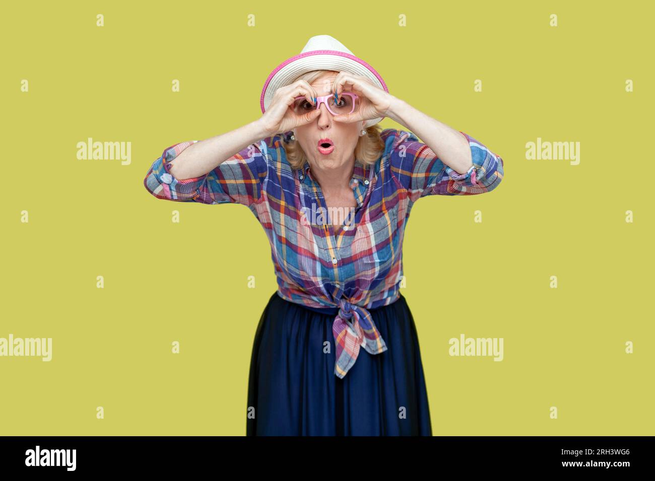 Senior woman makes binoculars gesture over eyes keeps mouth opened from wonder cannot believe in something awesome concentrated into distance. Indoor studio shot isolated on yellow background. Stock Photo