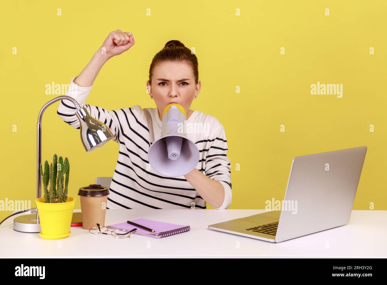 Portrait of angry woman loudly screaming at megaphone, making announce, protesting, wants to be heard, sitting on workplace. Indoor studio studio shot isolated on yellow background. Stock Photo