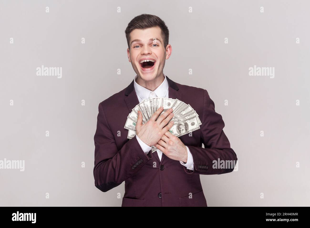 Portrait of extremely happy joyful handsome young man holding money, embracing dollar banknotes, smiling happily, wearing violet suit and white shirt. Indoor studio shot isolated on grey background. Stock Photo