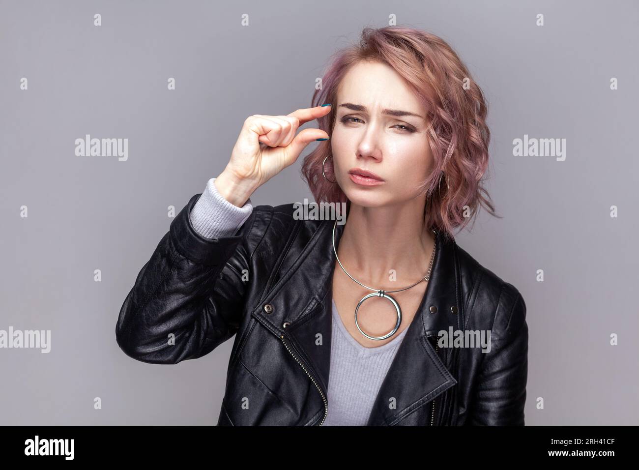 Portrait of attractive woman with short hairstyle standing looking at camera with frowning face, showing small gesture, wearing black leather jacket. Indoor studio shot isolated on grey background. Stock Photo