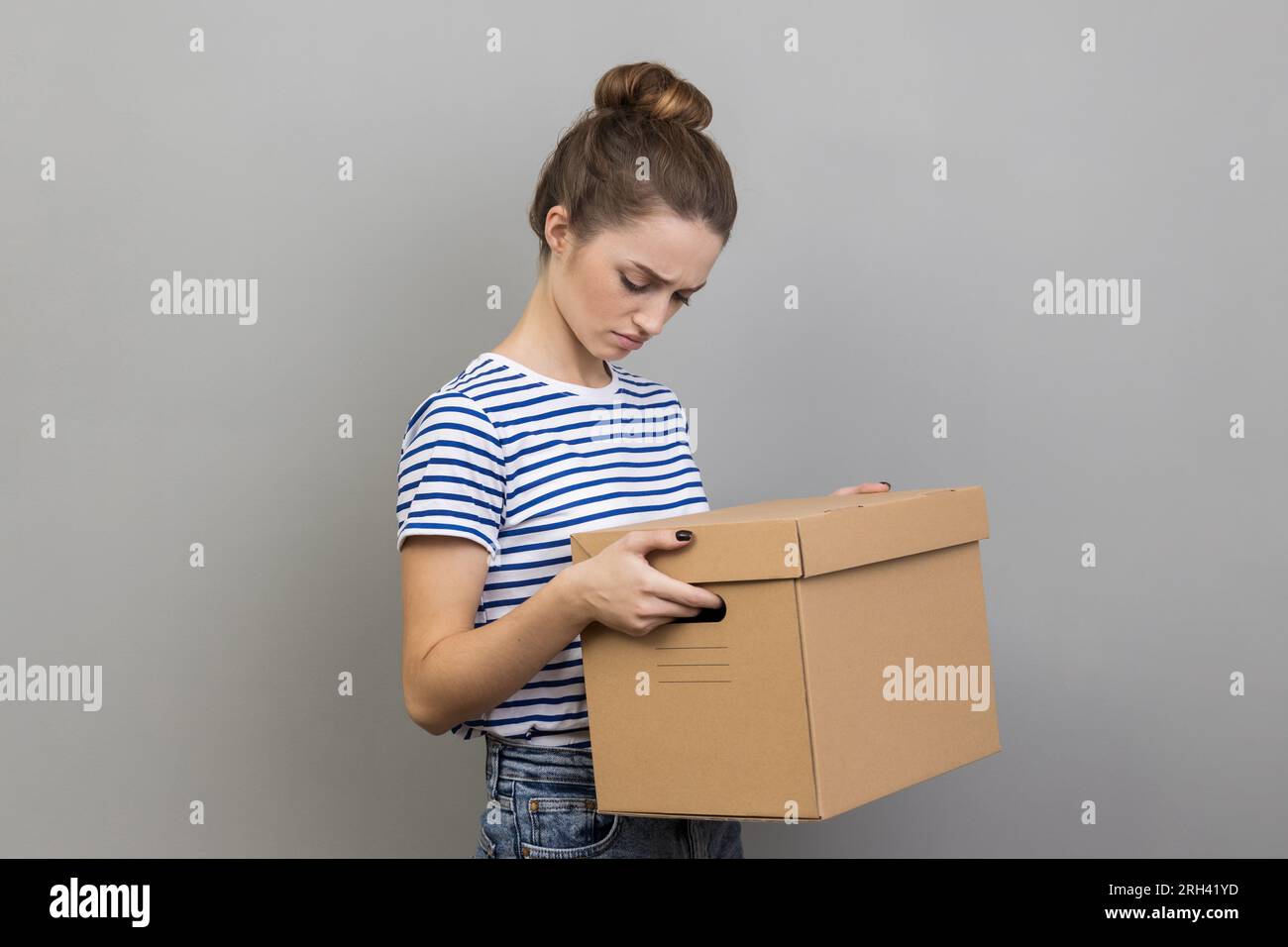 Portrait of sad upset attractive woman wearing striped T-shirt holding carton box with stuff, looking down with sorrow and sadness, lost her job. Indoor studio shot isolated on gray background Stock Photo