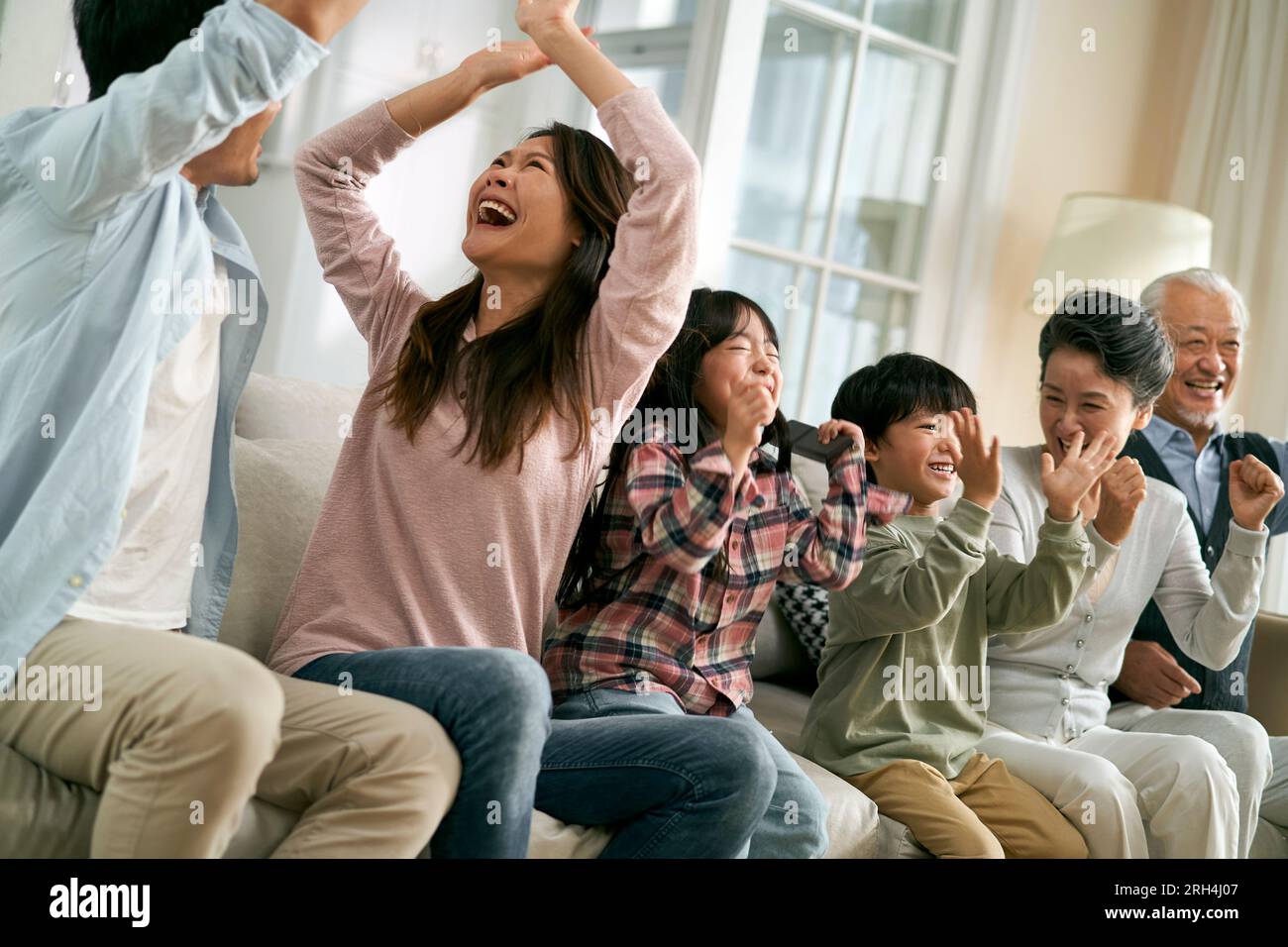 three generation asian family sitting on couch at home celebrating goal and victory while watching live broadcasting of football match Stock Photo