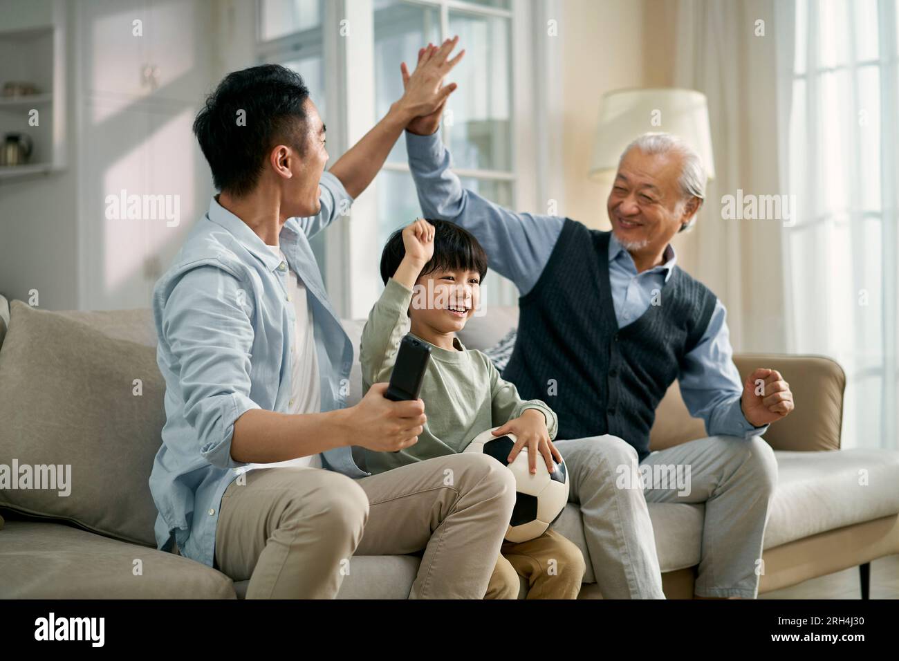 asian son father grandfather sitting on couch at home celebrating goal and victory while watching live broadcasting of football match on TV together Stock Photo