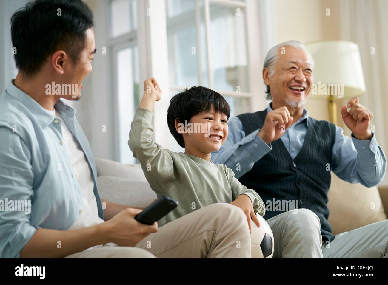 asian son father grandfather sitting on couch at home celebrating goal and victory while watching live broadcasting of football match on TV together Stock Photo