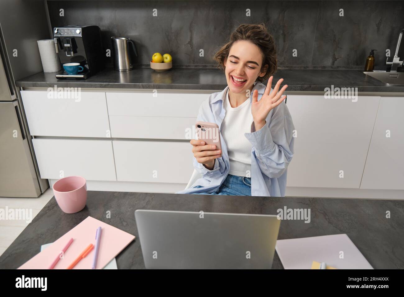 Chatty brunette woman sits at home, video chats with friend, waves hand at camera, holds smartphone and uses laptop Stock Photo