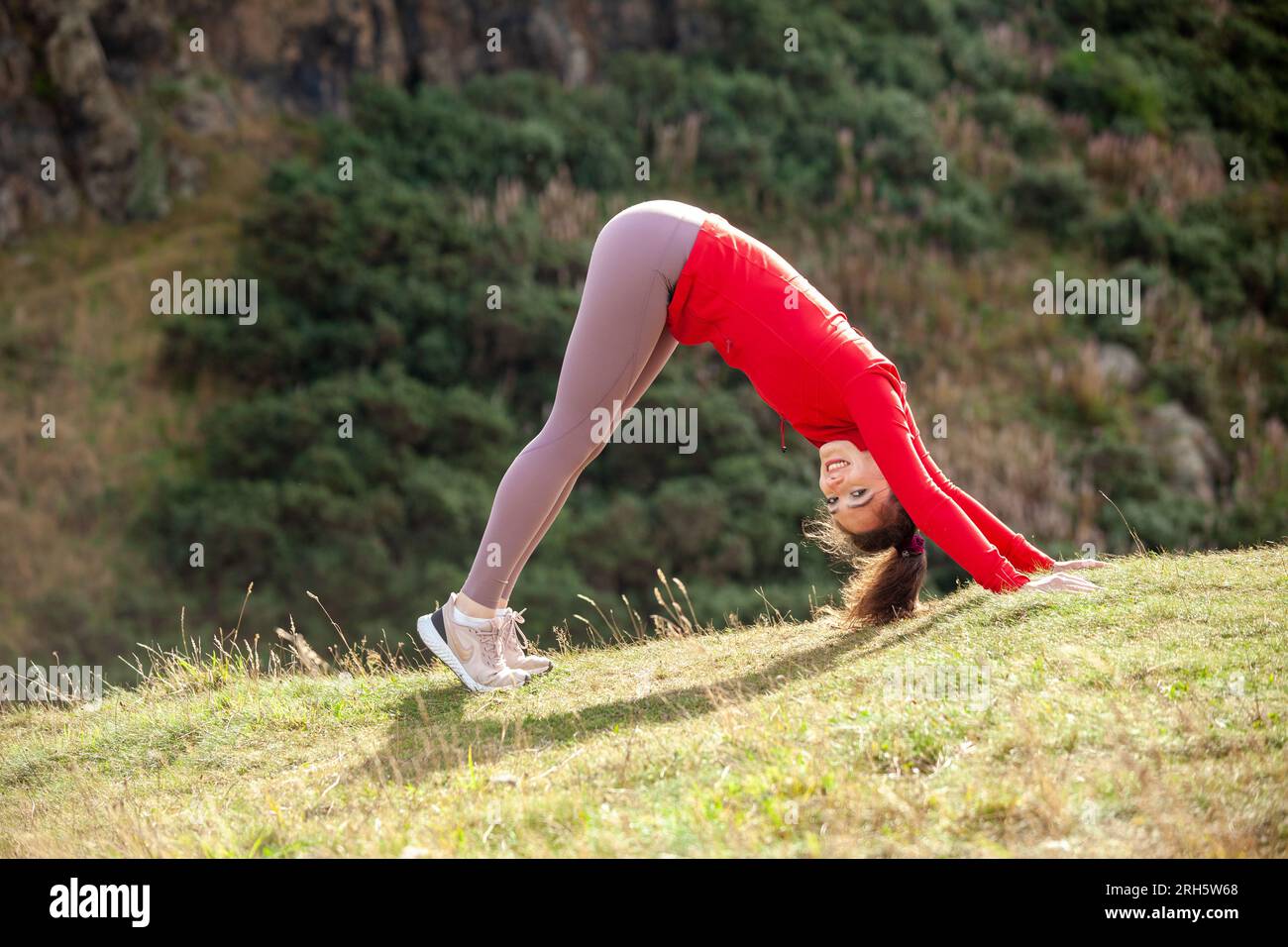 A young woman wearing leggings and performing the downward dog yoga stretch in a public park in Edinburgh Stock Photo