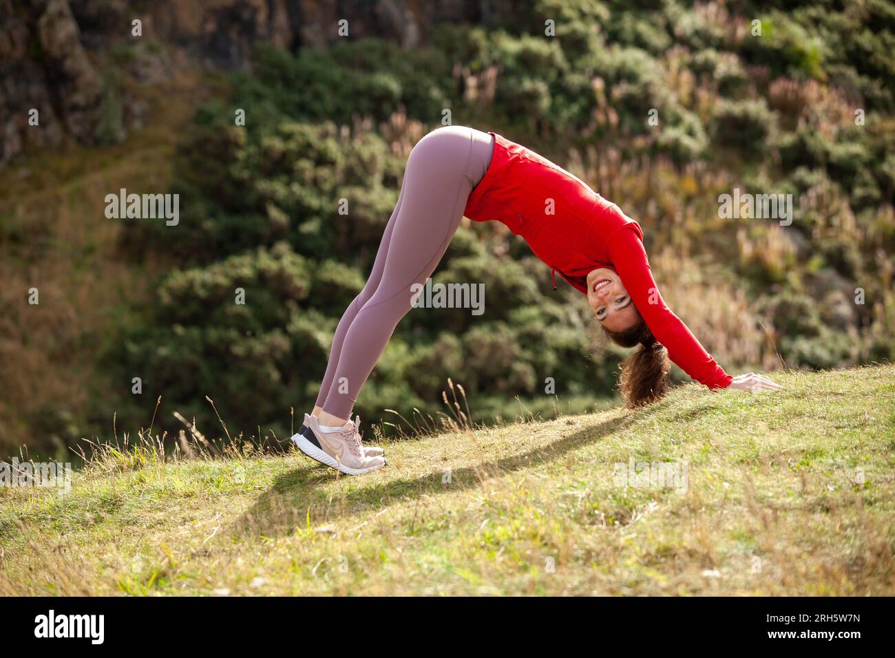 A young woman wearing leggings and performing the downward dog yoga stretch in a public park in Edinburgh Stock Photo