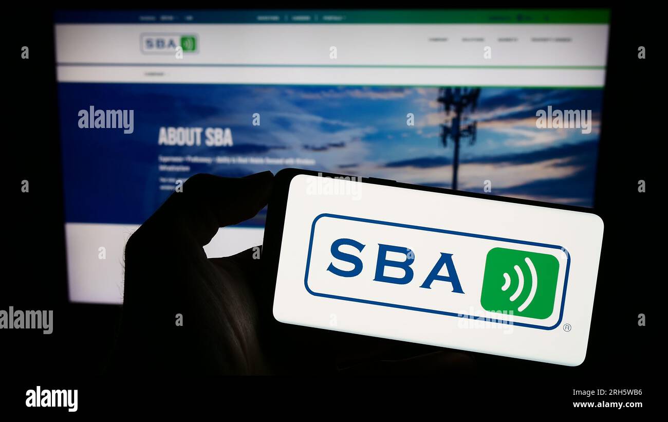 Person holding smartphone with logo of US company SBA Communications Corporation on screen in front of website. Focus on phone display. Stock Photo