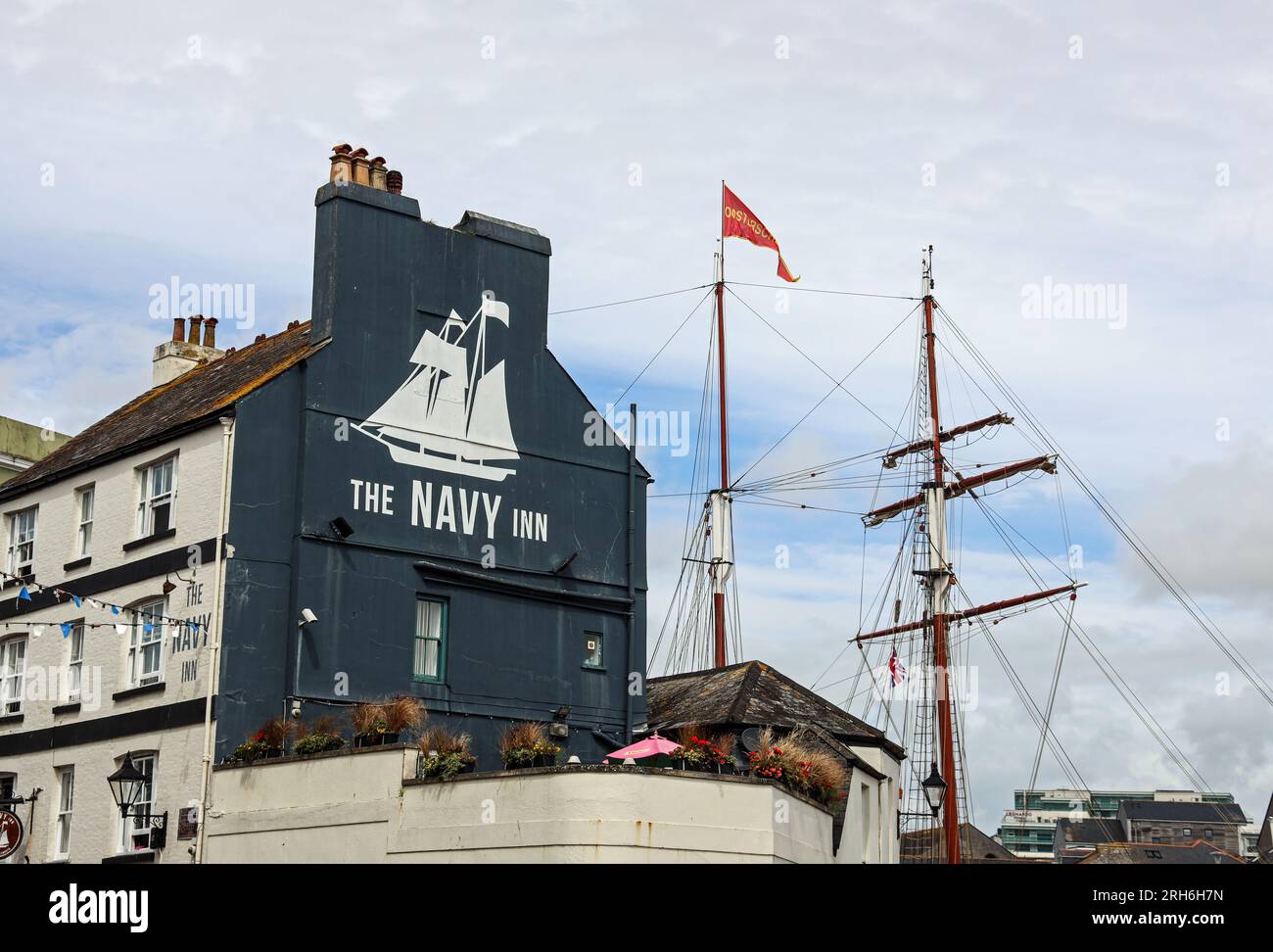 The side of the Navy Inn in Southside Street on Plymouth’s Barbican. Its logo above its rooftop beer garden reflected by the mast of the Oosterschelde Stock Photo