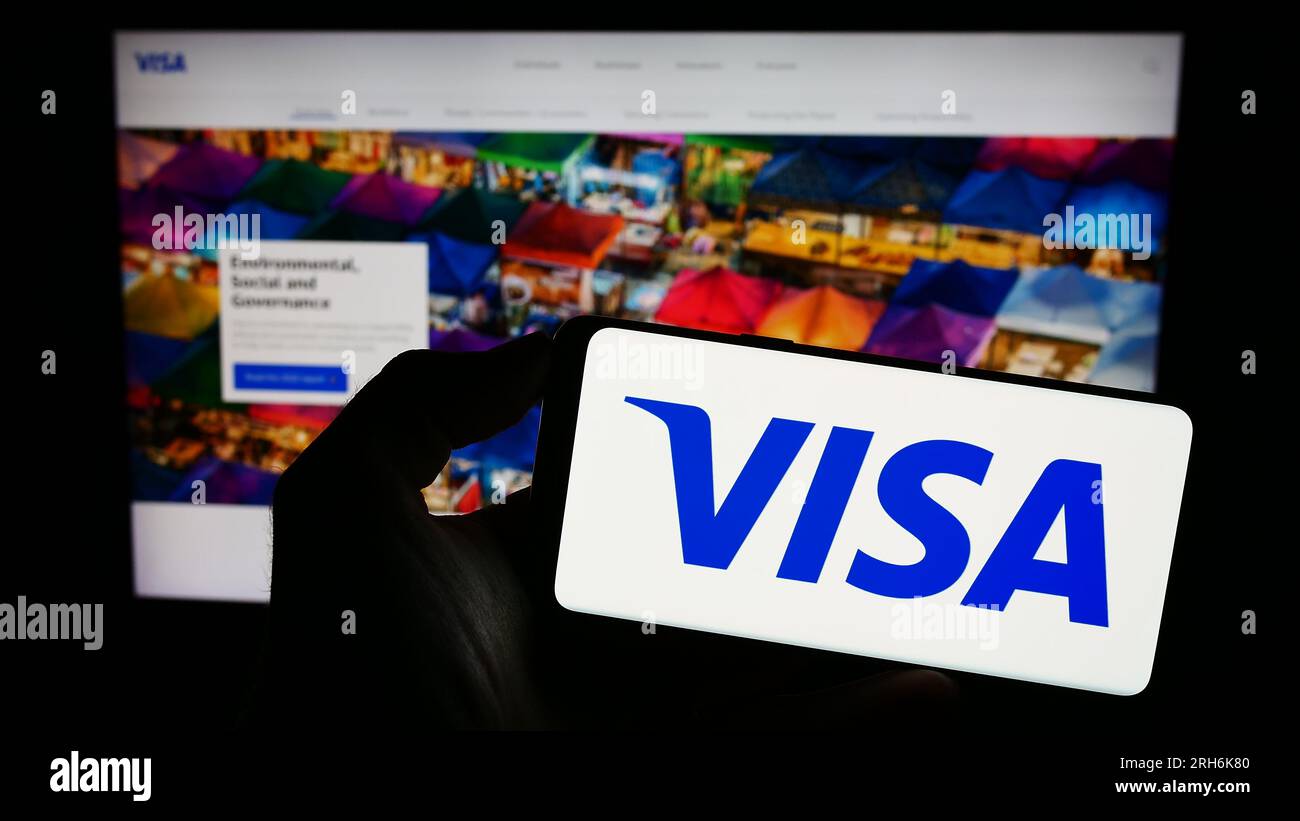 Person holding mobile phone with logo of American payment processing company Visa Inc. on screen in front of web page. Focus on phone display. Stock Photo