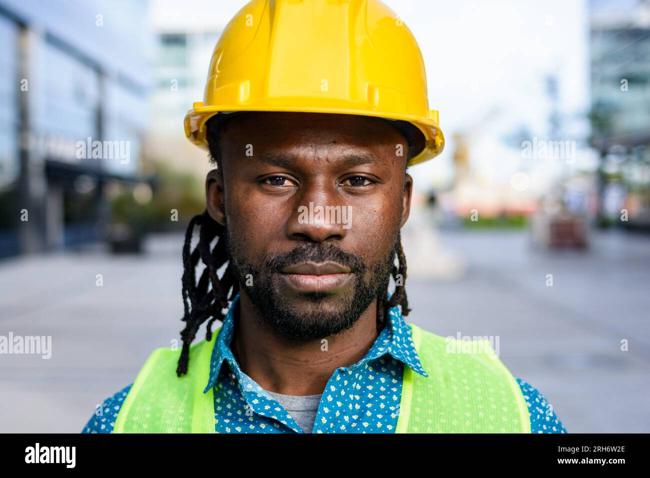 portrait of african ethnicity man with beard and dreadlocks, construction worker wearing helmet and reflective vest, standing outdoors, looking at cam Stock Photo