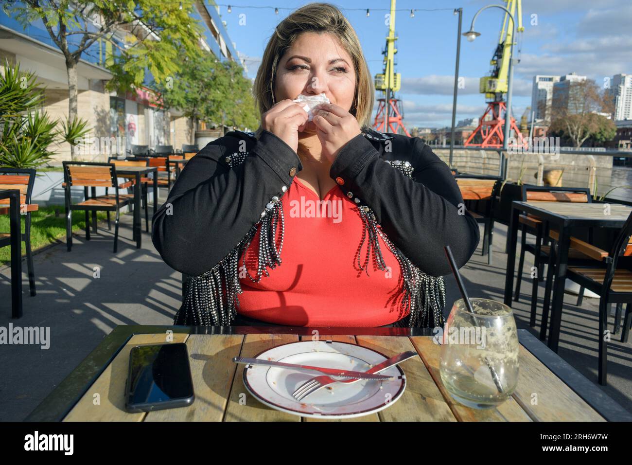 front view of young plus size latin woman sitting outside restaurant at sunset, she finished eating and is cleaning herself with napkin, concetp of pe Stock Photo