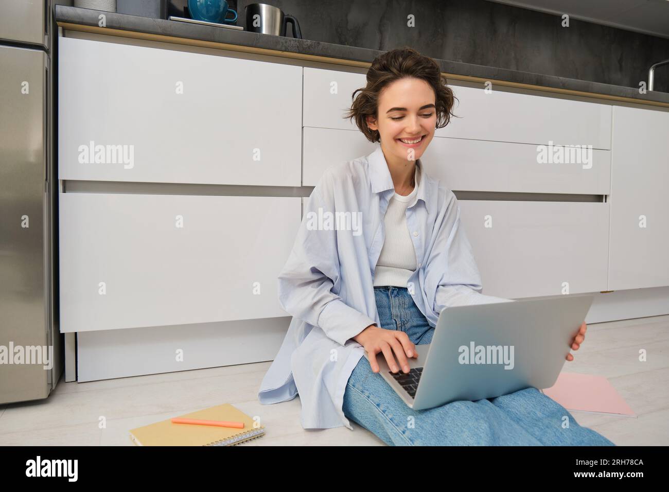 Portrait of young woman using laptop, sitting on kitchen floor, doing her homework, online chatting with friends on computer. Stock Photo