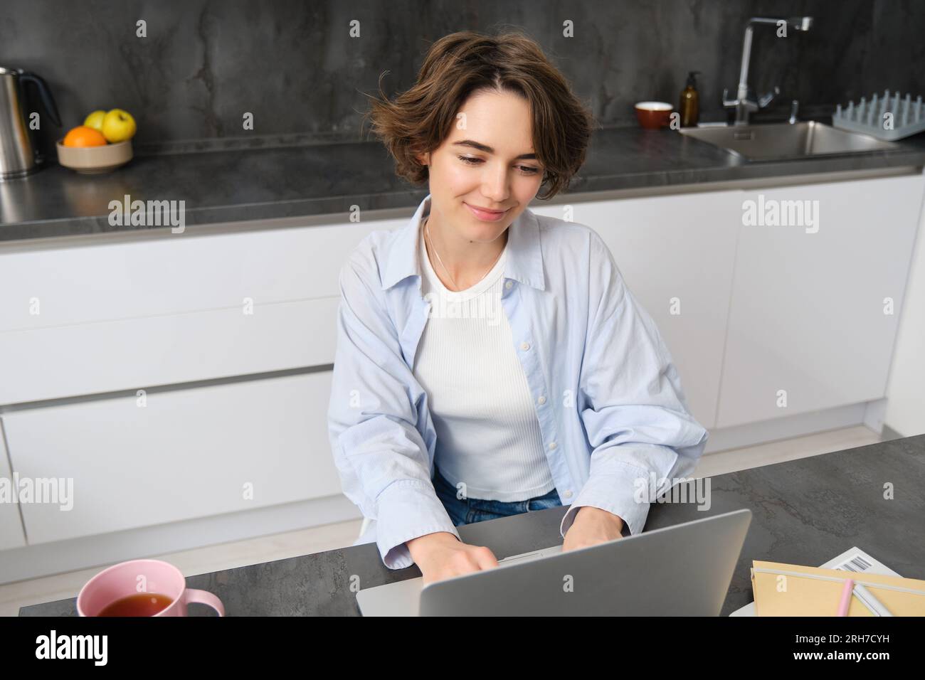 Young woman works from home, uses laptop in her kitchen. Student doing homework. Girl studies, watches online webinar, does distance learning. Courses Stock Photo