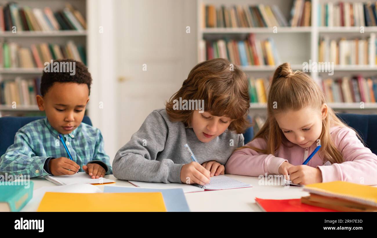 Attentive multiracial schoolkids boys and girl writing test, sitting at desk in classroom at primary school, panorama Stock Photo