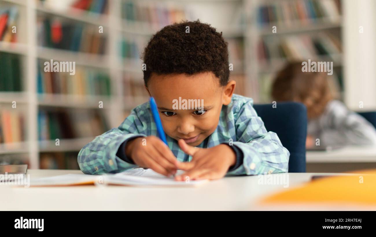 Concentrated black schoolboy sitting at desk in school and learning to write in copybook with pencil, studying, panorama Stock Photo