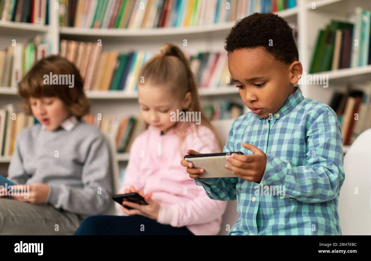 Diverse school kids students using smartphones in library, multiracial children holding mobile phones, playing games Stock Photo