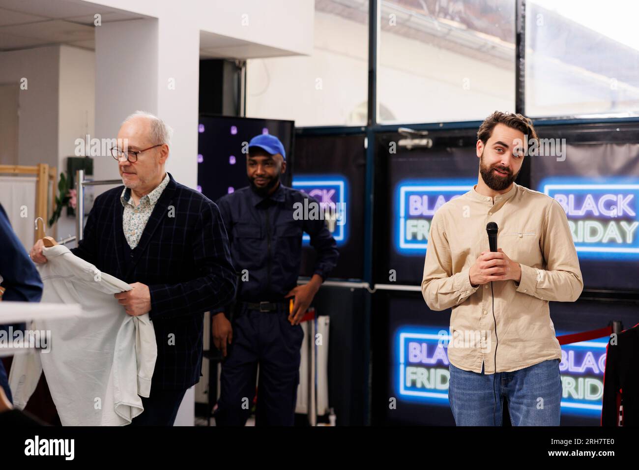 Young handsome smiling male TV news reporter talking into microphone reporting on crowd at local retail store during Black Friday sales. Male journalist making live broadcast about bargain-hunting Stock Photo