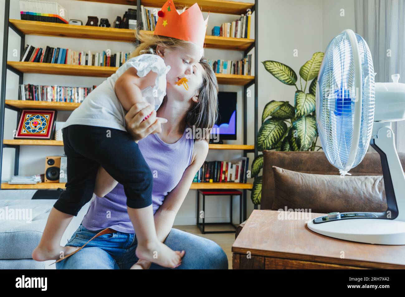 With the heat soaring, a wise mother and her daughter find respite and joy, dancing playfully in the cool breeze of a fan Stock Photo