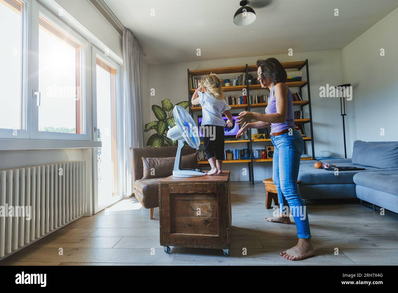 Mom, daughter find joy in fan's coolness, dancing and flying in the living room. Wise fun diverts from heat Stock Photo