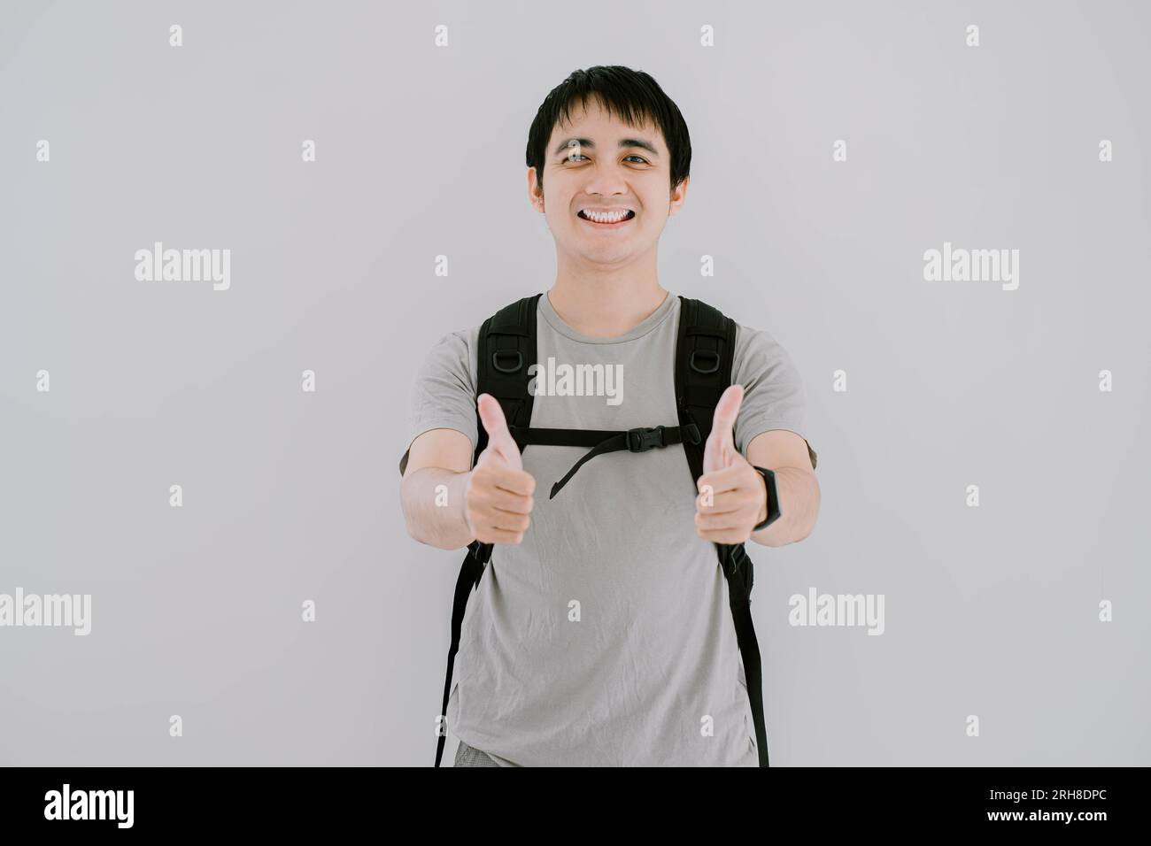Selective focus shot of young Asian man wearing sage green casual T-Shirt, smartwatch and backpack with smile expression is giving double thumb up Stock Photo
