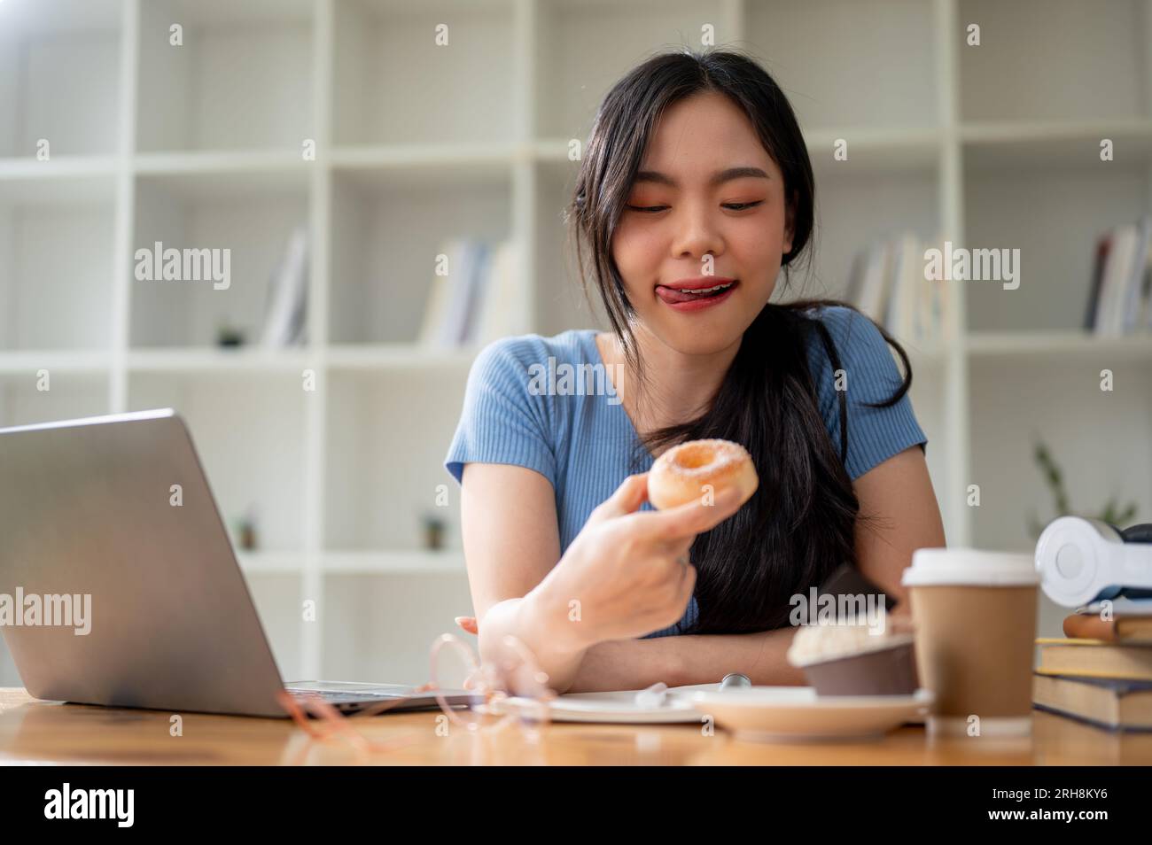 A pretty and happy young Asian woman enjoying her snack time, eating doughnuts at her study table at home. People and food concept Stock Photo