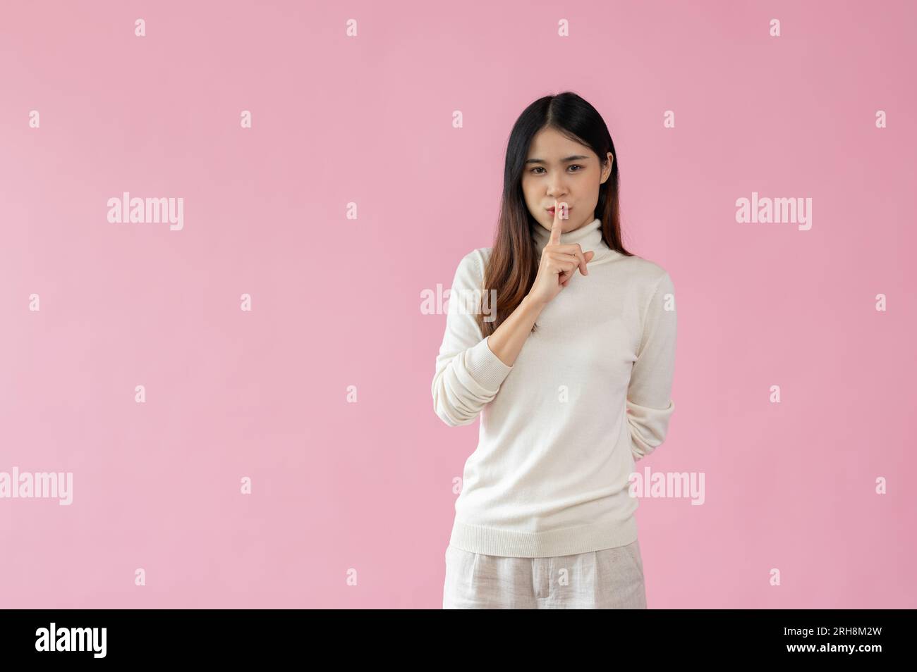 A beautiful Asian woman puts her finger on her lips, showing a shhh, keeping a secret, quiet, whisper, or silence gesture while standing against an is Stock Photo