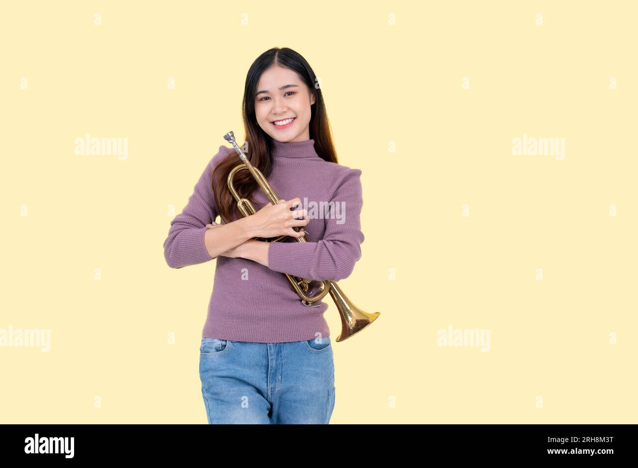 A smiling and pretty Asian woman in casual clothes stands against an isolated yellow background with a trumpet in her hands. Musician, creative hobby, Stock Photo