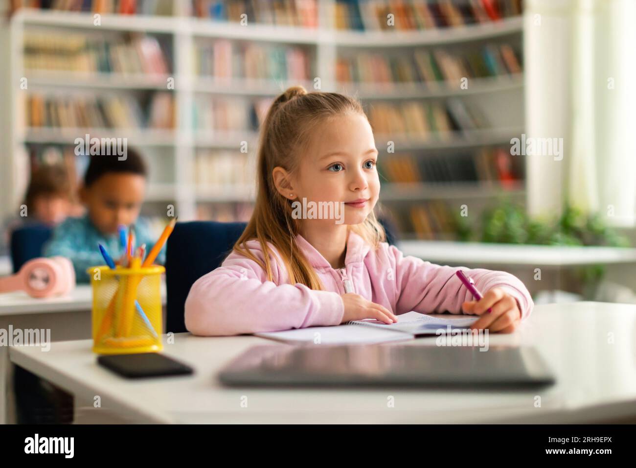 Academic concept. Smart caucasian school girl sitting at desk in classroom, listening teacher and writing in notebook Stock Photo