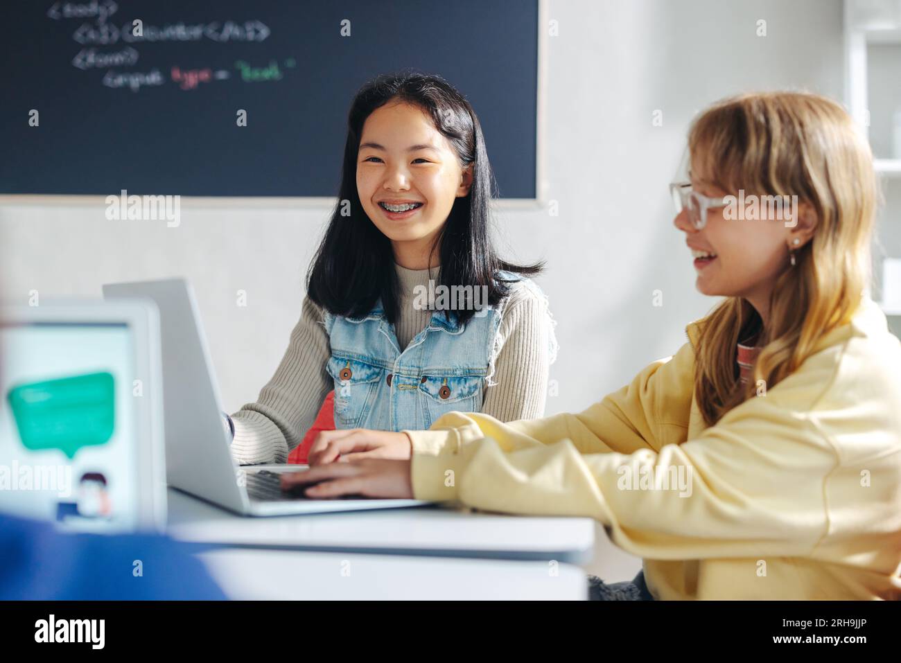 Elementary school students learning coding and computer programming in a classroom. Two young girls smiling as they sit next to each other, using a la Stock Photo