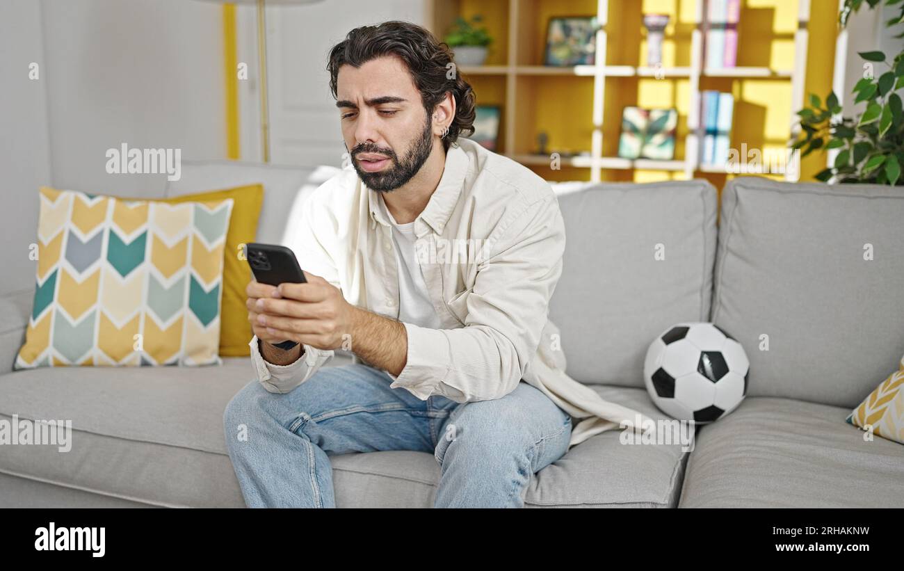 Young hispanic man watching soccer match using smartphone at home Stock Photo